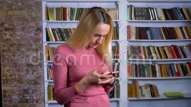 金发白种人年轻女孩在书架背景上专心看智能手机的肖像。